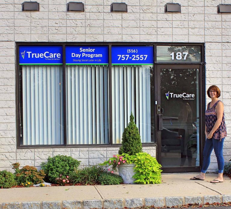 The TrueCare storefront at 187 Veterans Boulevard, with TrueCare, Senior Day Program and (516) 757-2557 signs in the windows and Carol, TrueCare’s owner, standing beside the entrance