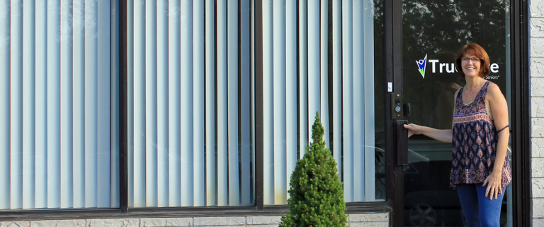 Carol with her hand on the handle of TrueCare’s glass entrance door, with the TrueCare logo on the glass and a small evergreen shrub by the windows