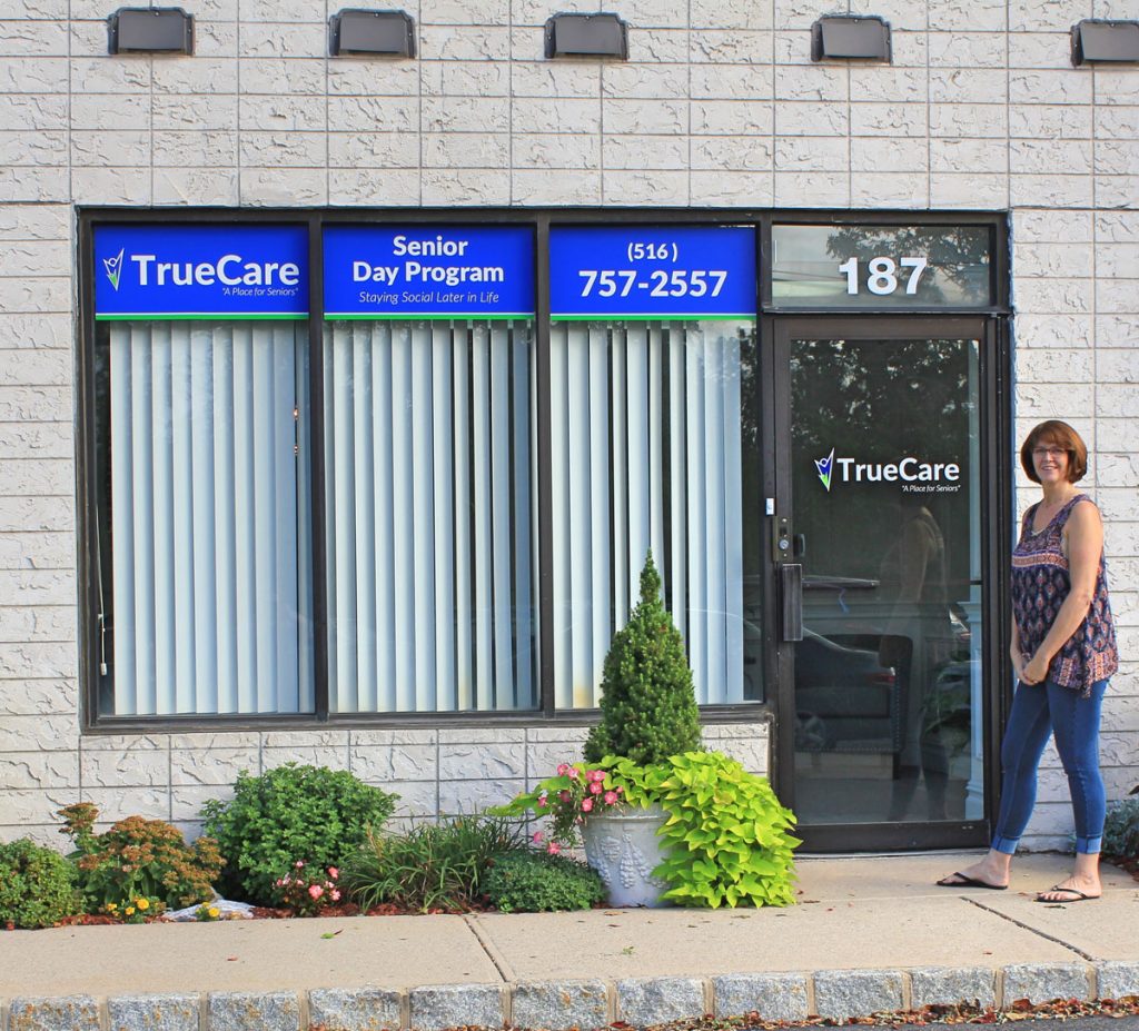 The TrueCare storefront at 187 Veterans Boulevard, with blue TrueCare, Senior Day Program and phone-number signs in the windows and Carol, TrueCare’s owner, standing beside the glass entrance door