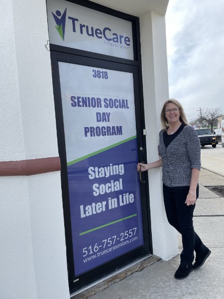 Carol at the TrueCare entrance at 3818 Merrick Road, with a Senior Social Day Program sign on the door