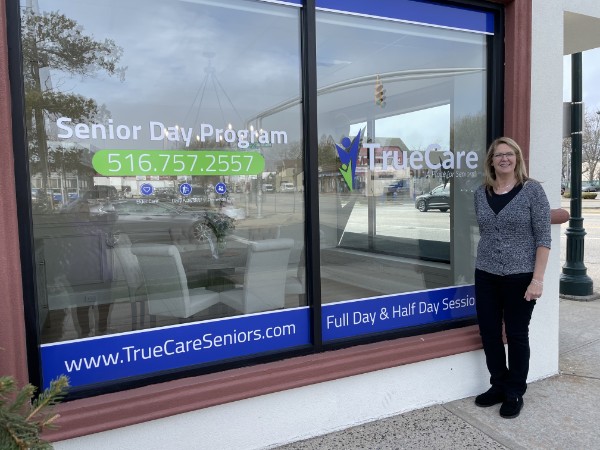 The TrueCare storefront on Merrick Road in Seaford, with Senior Day Program, 516.757.2557 and TrueCare signs on the windows, and Carol, TrueCare’s owner, standing beside it